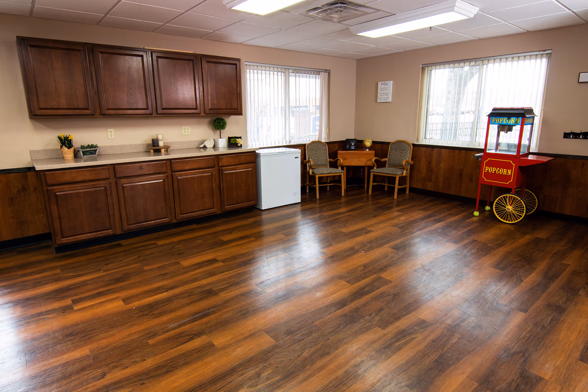 A room with wooden flooring and beige walls featuring a kitchenette with wooden cabinets, a small white refrigerator, two chairs with a small wooden table between them, and a red popcorn machine near a window with vertical blinds.