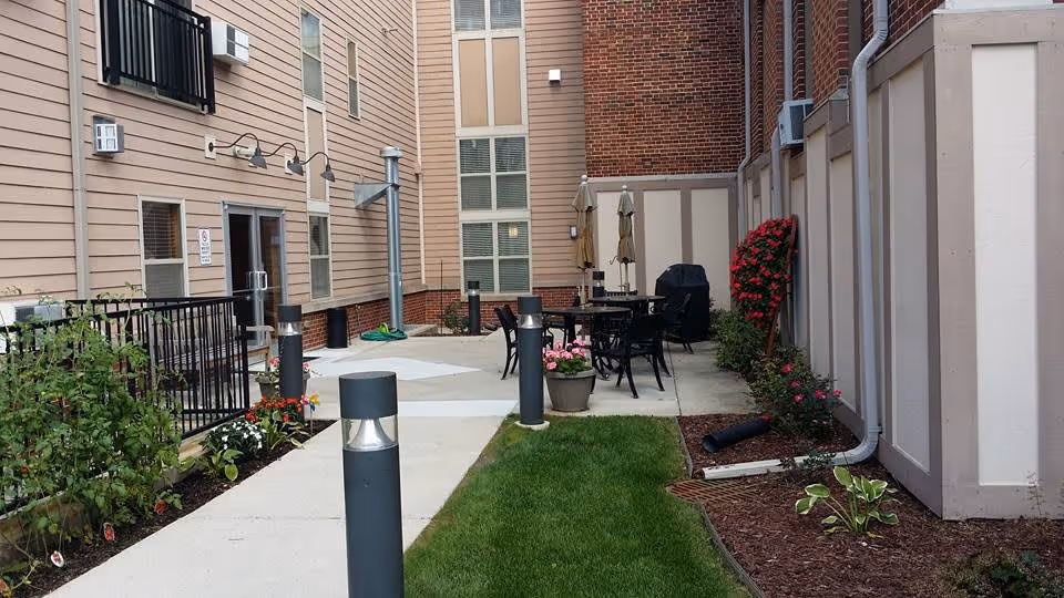 Outdoor patio area between buildings with a concrete walkway, green grass, flower beds, black metal table and chairs, umbrellas, and a barbecue grill.