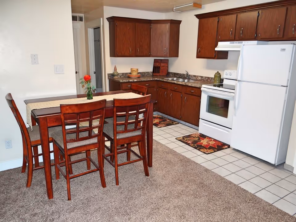 A kitchen and dining area with wooden cabinets, a white refrigerator, white stove, and a dark wooden dining table with four matching chairs. The floor in the kitchen area is tiled, while the dining area has carpet. A single red flower in a small green vase is placed on the dining table.