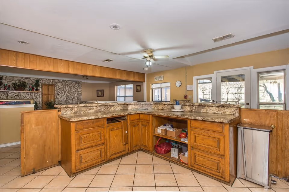 Wooden L-shaped service counter with marble-look countertops, open shelving and tile floor in a communal kitchen area.