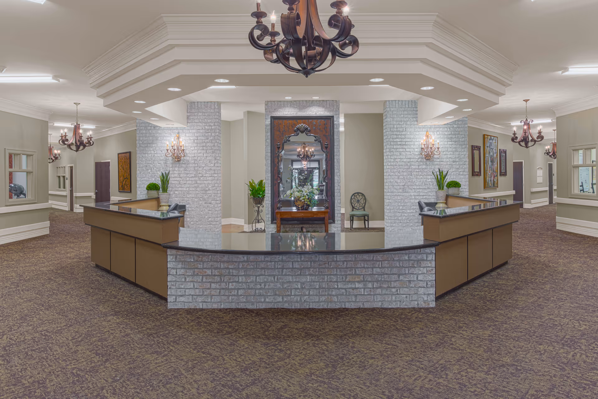 Spacious interior lobby area with a large central reception desk made of brick and wood, decorated with plants and a large ornate mirror on the back wall. The space features carpeted floors, multiple chandeliers, wall sconces, and neutral-colored walls with artwork and windows on the sides.