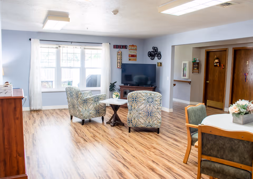 A bright and cozy living room area in a senior living facility with two patterned armchairs facing a flat-screen TV on a wooden stand. The room has light blue walls, large windows with white curtains letting in natural light, and wood flooring. There is a small table between the chairs with a flower arrangement, and a round dining table with chairs and a flower centerpiece is partially visible. Wooden doors and wall decorations are seen in the background.