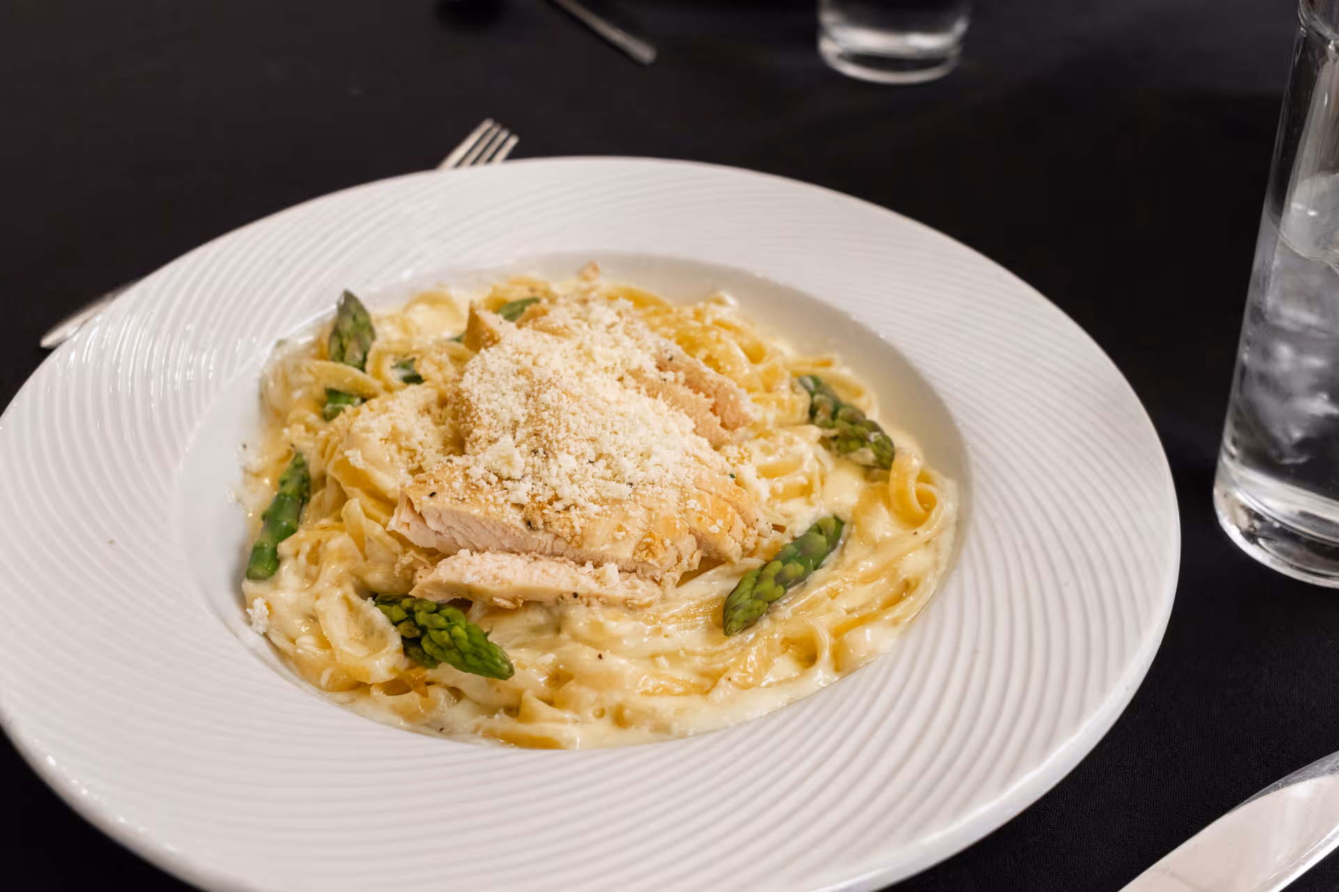 A white plate with creamy fettuccine pasta topped with sliced chicken breast, asparagus spears, and grated cheese, placed on a black tablecloth with a glass of water and silverware nearby.