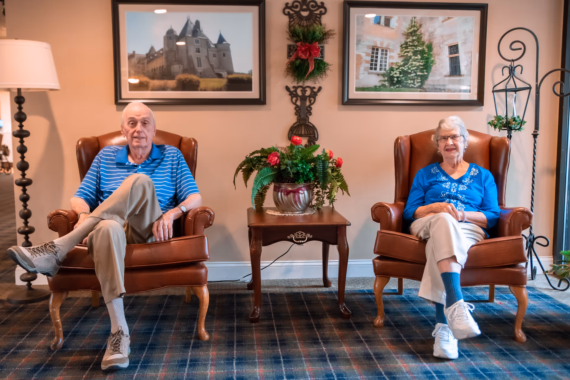 An elderly man and woman sitting in brown leather armchairs in a cozy living room area. Between them is a wooden side table with a decorative plant. The wall behind them features two framed pictures and a small wreath with a red bow. The floor is carpeted with a plaid pattern.