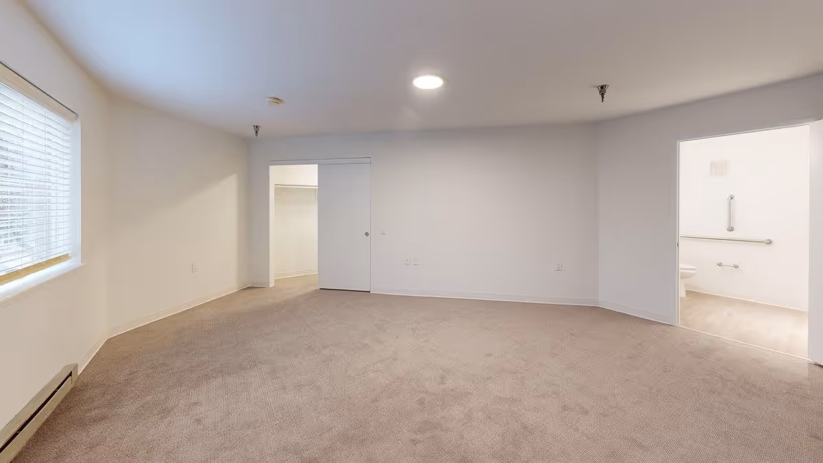 Empty room with beige carpet and white walls, a window with blinds on the left, a closet with a sliding door in the center, and an open doorway on the right leading to a bathroom with grab bars and a toilet.