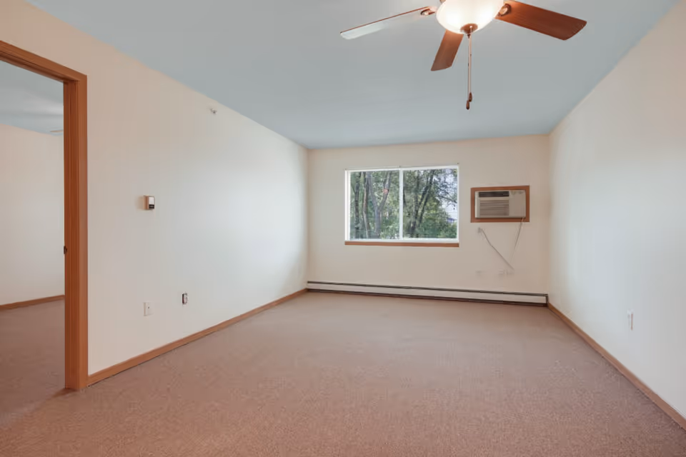 Empty room with beige walls and carpet, a window with a view of trees, a ceiling fan with wooden blades, and a wall-mounted air conditioning unit. There is an open doorway leading to another room with similar carpet and wall color.