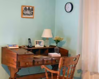 Wooden desk with drawers, chair, lamp, photos, and clock in teal-walled room.