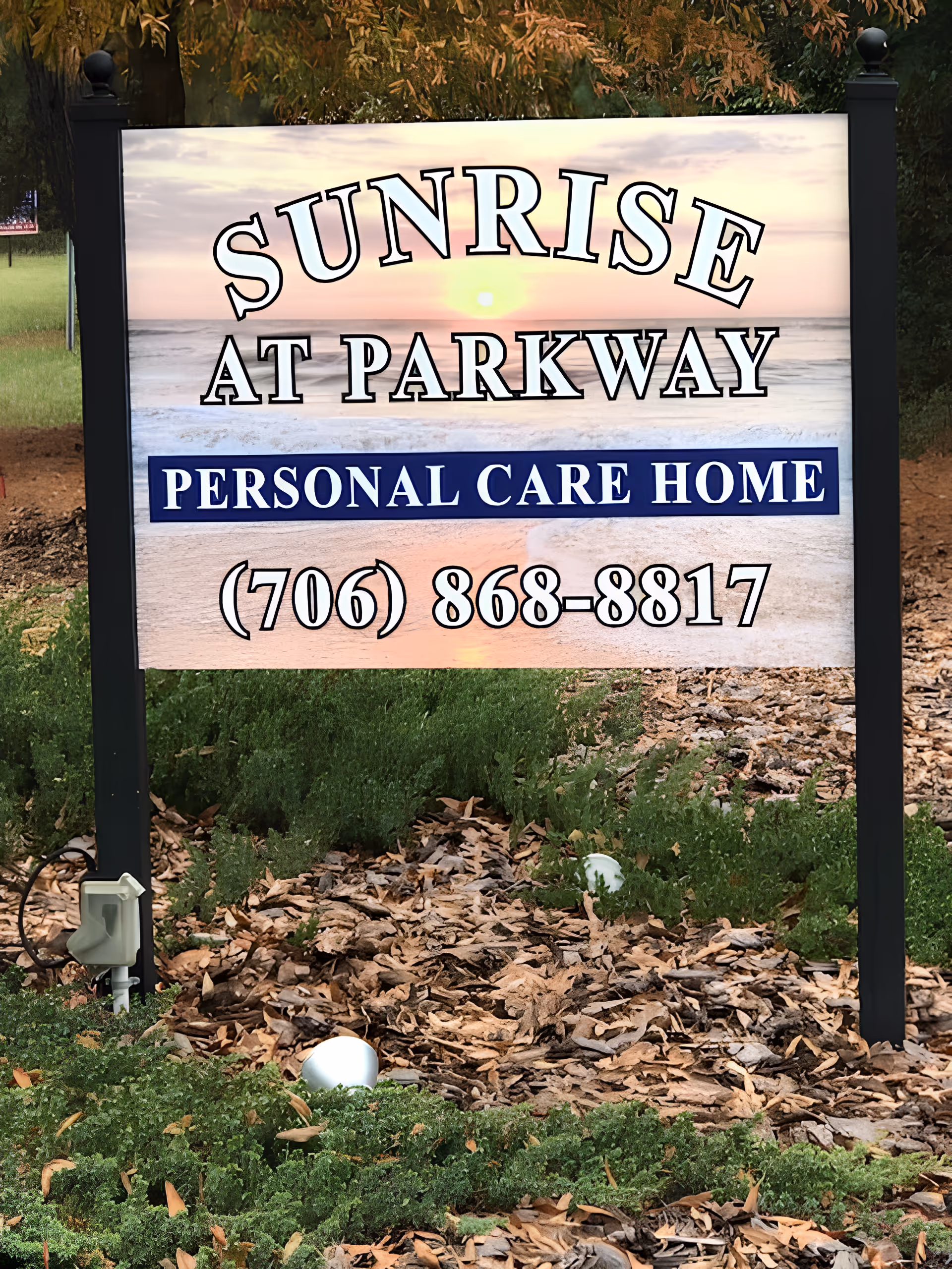 Outdoor sign for Sunrise At Parkway Personal Care Home with a phone number (706) 868-8817, set against a background image of a sunrise over the ocean. The sign is mounted on black posts and surrounded by green plants and fallen leaves.