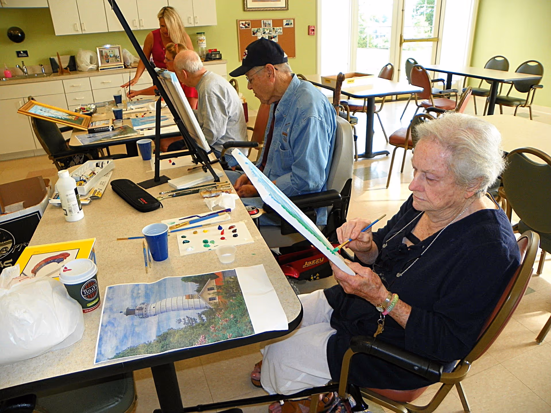A group of elderly individuals seated around a table in a well-lit room, engaged in painting activities. They have canvases, paint palettes, and brushes, with one woman in the foreground focused on her painting. The room has large windows, tables, chairs, and a kitchenette area in the background.