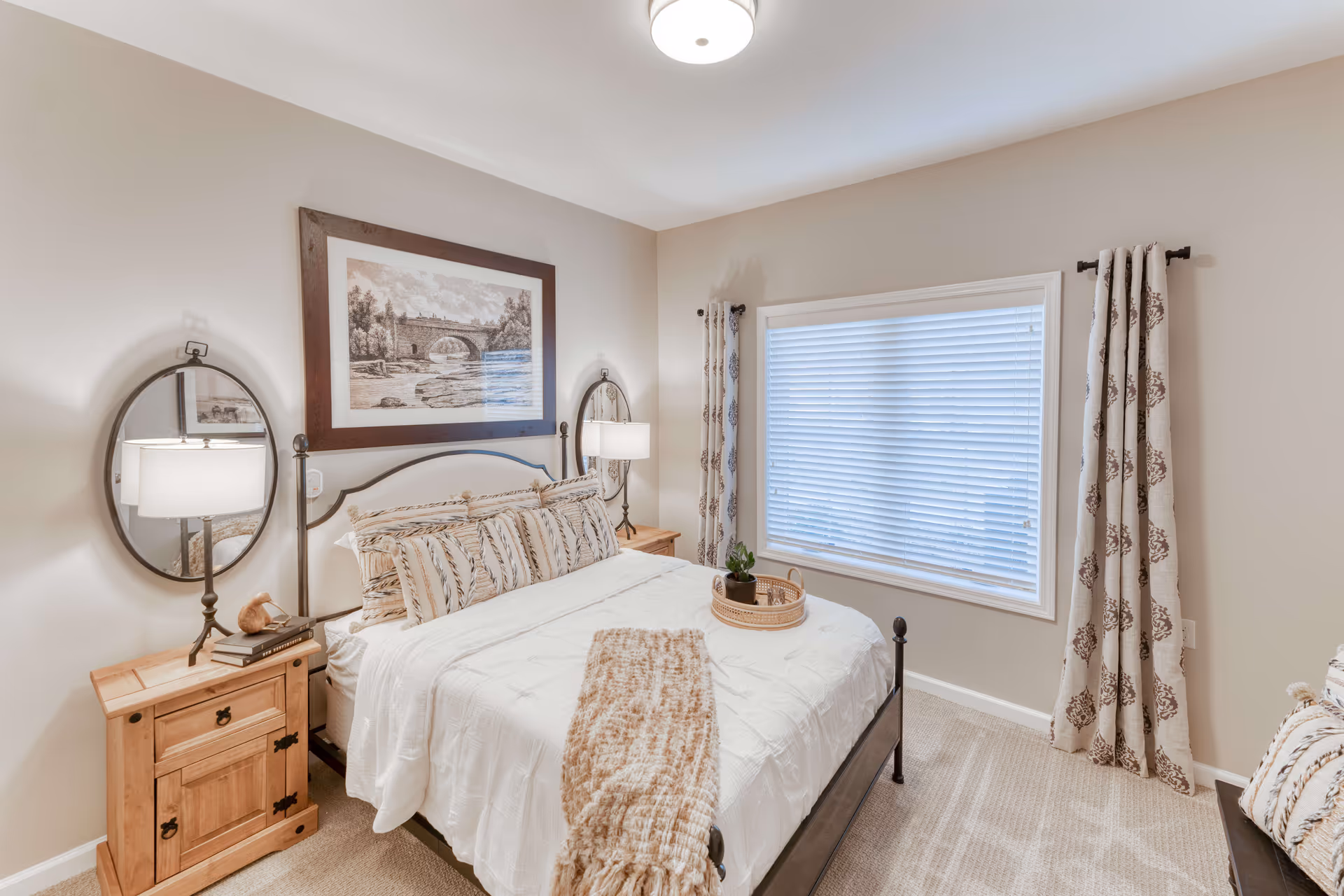A cozy bedroom with a neatly made bed featuring white bedding and patterned pillows. There are two wooden nightstands on either side of the bed, each with a lamp and an oval mirror mounted on the wall above. A framed picture of a stone bridge over a river hangs above the bed. The room has beige walls, a large window with white blinds and patterned curtains, and a beige carpeted floor.