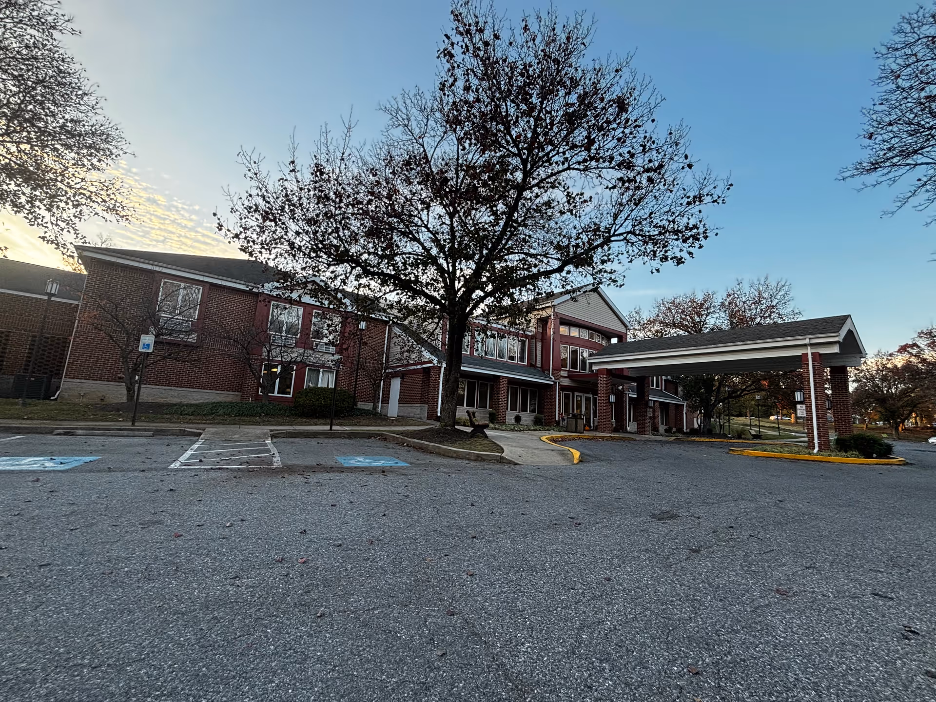Exterior view of a two-story brick building with red accents, a covered entrance, and a parking lot with handicap parking spaces. Trees with sparse leaves are visible around the building under a clear sky.
