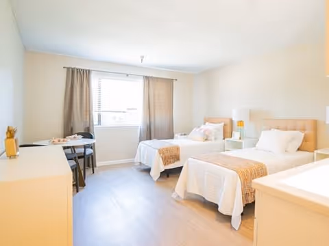 A bright and clean bedroom with two single beds, each with white bedding and a beige throw blanket. There is a small white nightstand with a lamp between the beds. A window with beige curtains lets in natural light. A small table with two chairs is placed near the window. The room has light-colored walls and wooden flooring.