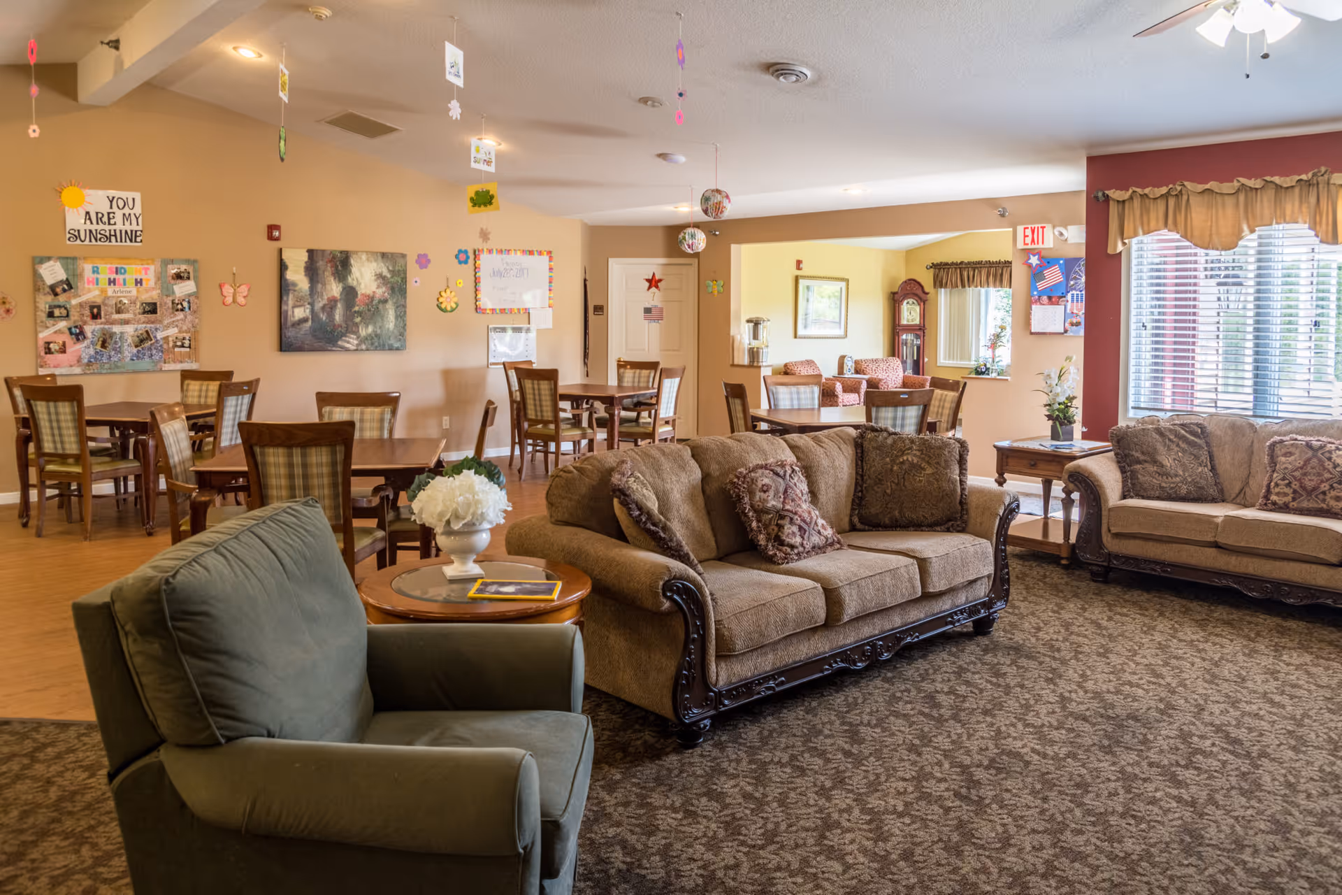 A cozy senior living common area with two beige sofas and a green armchair arranged around a small wooden table with a white flower vase. In the background, there are several wooden dining tables and chairs, bulletin boards with decorations and photos, and a window with blinds and curtains letting in natural light.