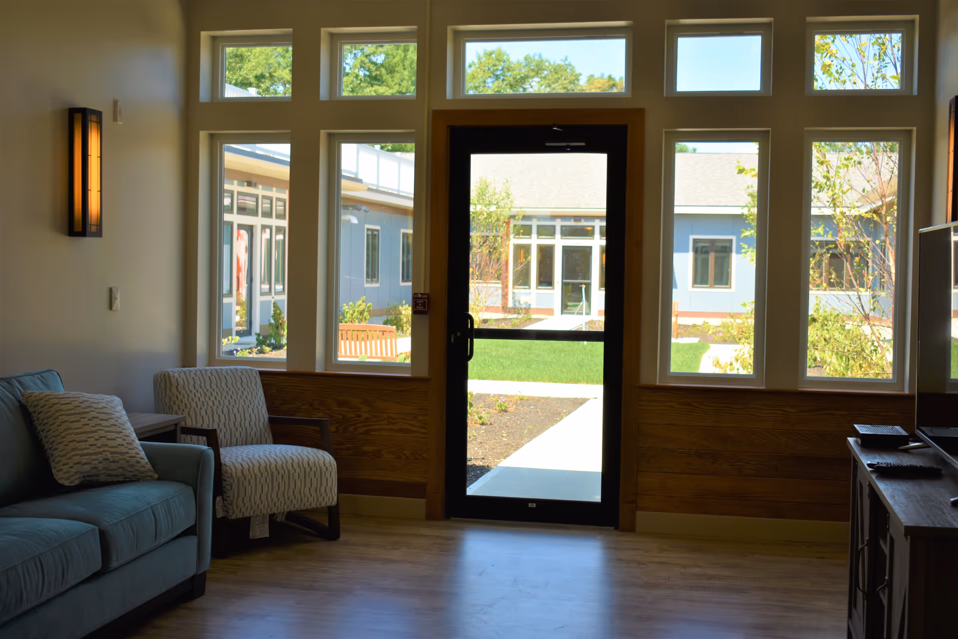 A cozy sitting area inside Harmony Homes By The Bay featuring a blue sofa with a patterned pillow, a cushioned armchair, and a wooden side table. Large windows and a glass door provide a view of a green courtyard with benches and a blue building exterior.