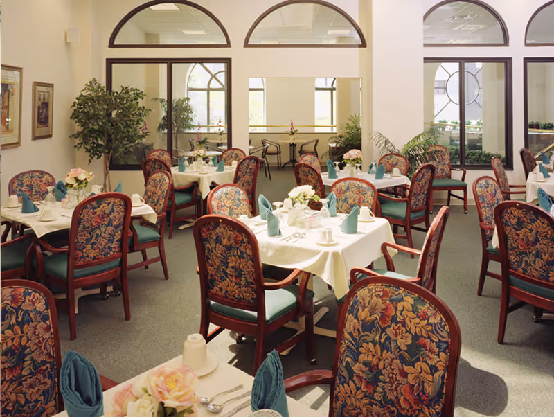 A dining room in Amber Court Assisted Living in Elizabeth with multiple tables covered in white tablecloths, each set with teal folded napkins, cups, and floral centerpieces. The chairs have wooden frames with floral-patterned upholstery. Large windows and mirrors with arched tops line the walls, and there are several potted plants around the room.