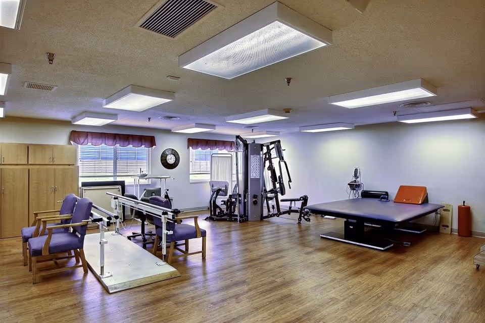 A spacious physical therapy or rehabilitation room with wooden flooring, equipped with parallel bars for walking exercises, several purple cushioned chairs, a multi-functional exercise machine, and a large padded therapy table. The room has two windows with purple valances, a wall clock, and bright ceiling lights.