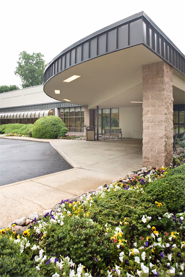 Covered entrance canopy with stone pillars, a concrete driveway and landscaped flower beds in front of a building.