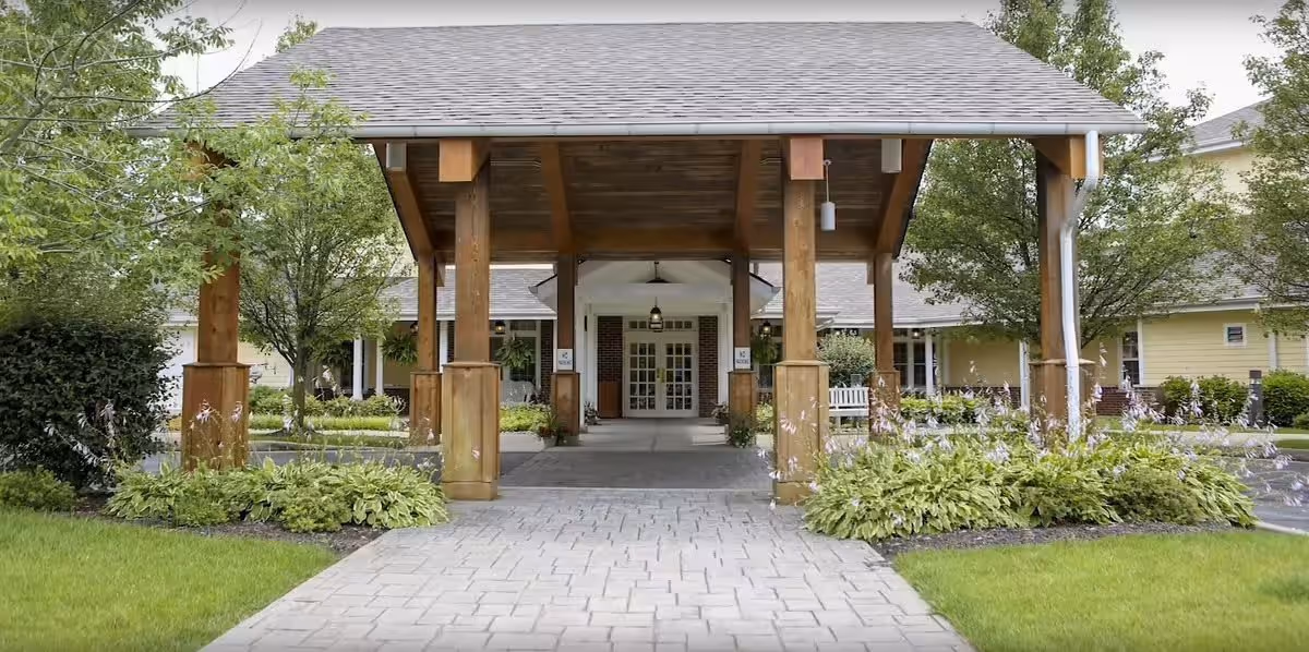 Entrance of River Terrace Retirement Community featuring a covered wooden canopy supported by large wooden pillars, with a paved walkway leading to double glass doors. The area is surrounded by green shrubs, trees, and well-maintained landscaping.