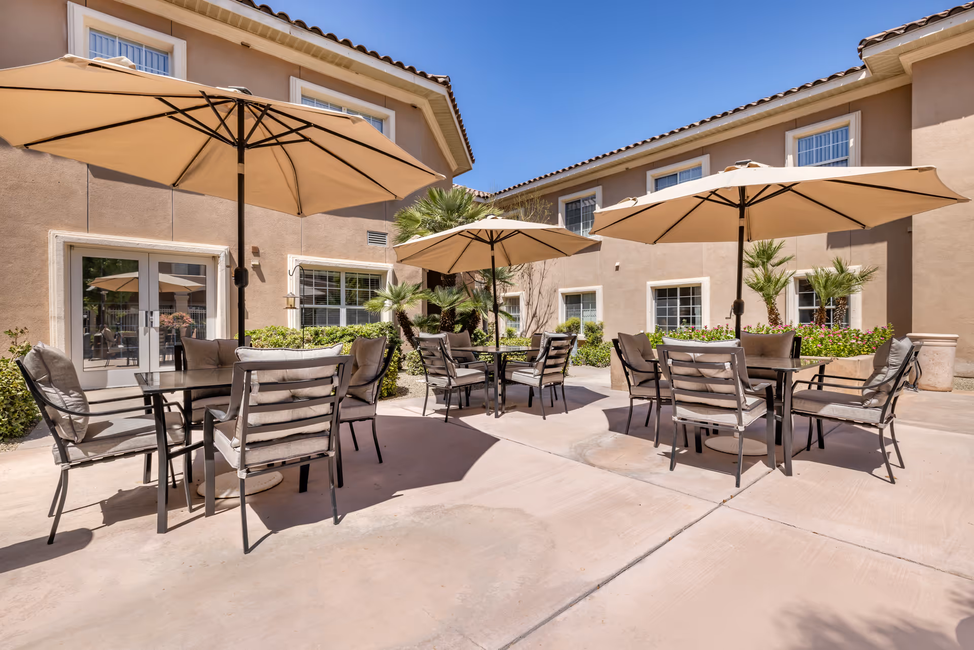 Outdoor patio area with multiple tables and cushioned chairs under large beige umbrellas, surrounded by a beige building with windows and some greenery including palm plants and flowers.