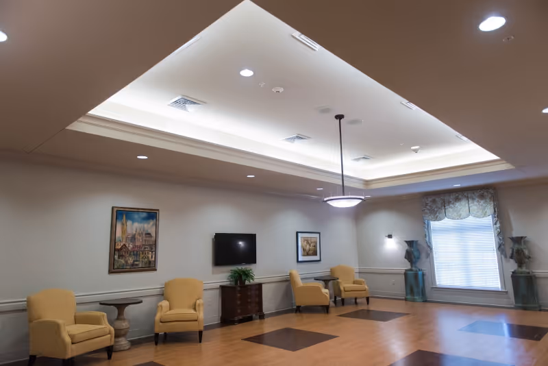A spacious, well-lit common area with a tray ceiling featuring recessed lighting and a central hanging light fixture. The room has light-colored walls adorned with framed artwork and a flat-screen TV mounted on one wall. There are several yellow upholstered armchairs arranged along the walls, small tables, and decorative vases near a window with blinds and a valance. The floor is a mix of wood and darker square insets.