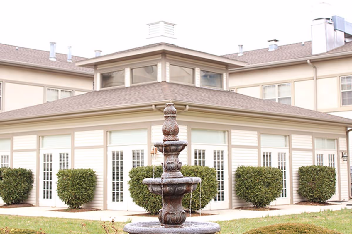 Exterior view of a senior living facility building with a tiered stone fountain in the foreground and trimmed shrubs along the windows.