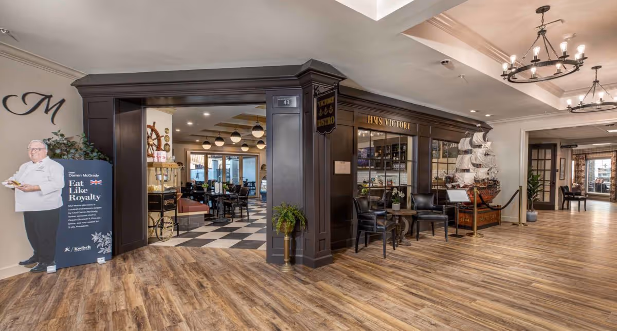 Interior view of a senior living facility showing the entrance to HMS Victory Bistro with wooden floors, dark wood paneling, a model ship, and seating area with two chairs and a small table. To the left, there is a life-size cutout of a chef with a sign promoting dining services. The hallway extends to the right with additional seating visible in the distance.