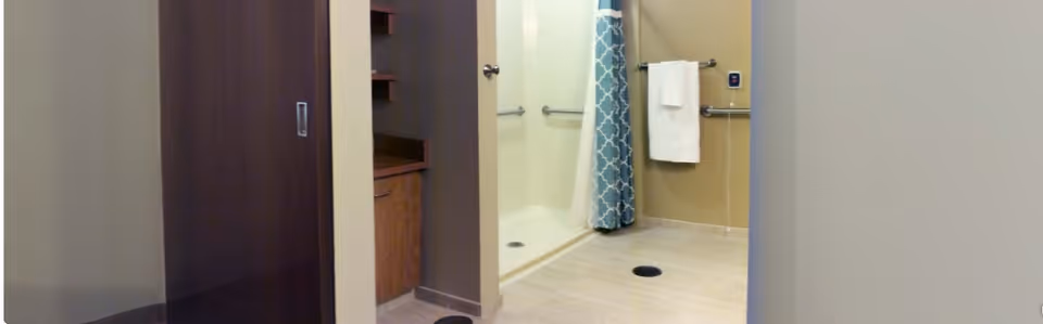 A bathroom with a walk-in shower featuring a blue and white patterned shower curtain, grab bars on the walls, a towel hanging on a bar, and a wooden cabinet with a countertop visible to the left.