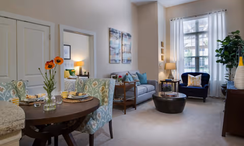 A cozy senior living apartment interior featuring a small round dining table set for two with floral-patterned chairs, a living area with a gray sofa, a dark blue armchair, a round coffee table, and a tall window with white curtains letting in natural light. A bedroom is partially visible through an open doorway, and decorative artwork and plants add warmth to the space.