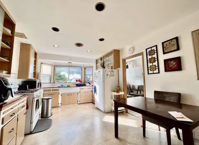 A bright kitchen with beige cabinets, a white refrigerator, a stove, a microwave, and a trash can. There is a dark wooden dining table with a chair on the right side, and decorative wall art above it. Large windows above the sink let in natural light, and a doorway leads to another room with white cabinets and chairs.