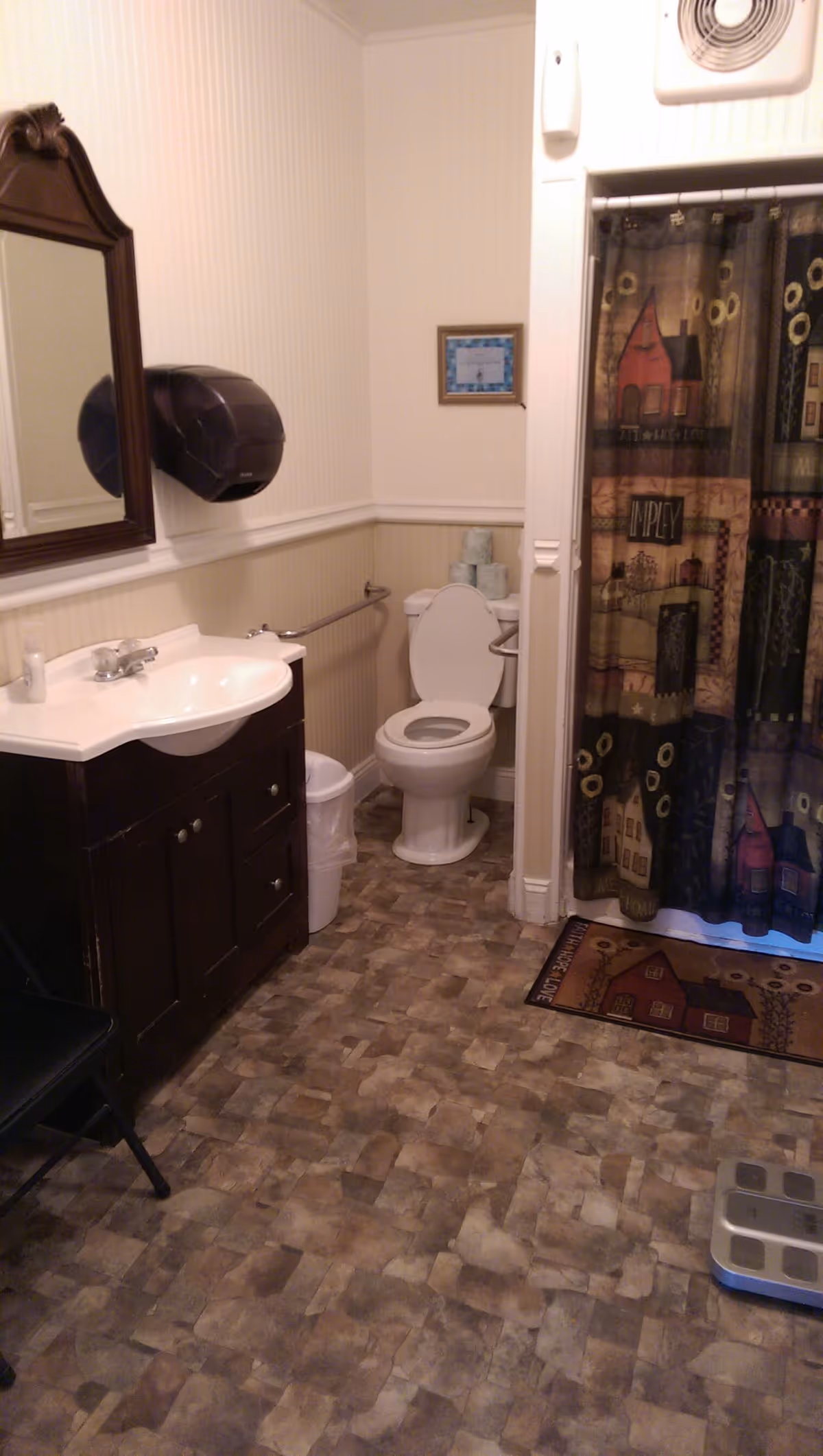 A bathroom with a white toilet, a dark wood vanity with a white sink, a mirror above the sink, a wall-mounted paper towel dispenser, a shower with a decorative curtain featuring houses, a small framed picture on the wall, a trash can, and a floor scale on the tiled floor.
