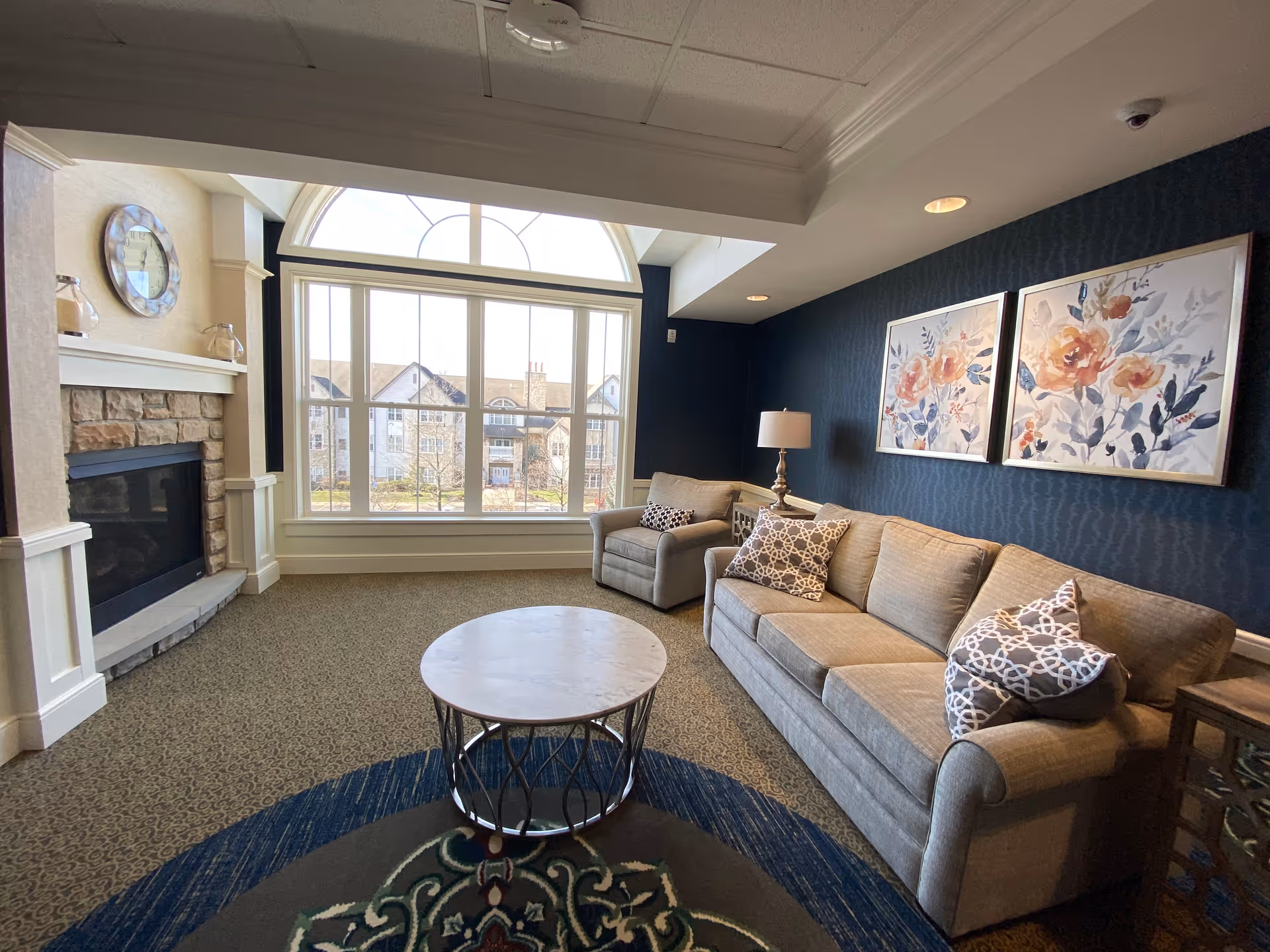 A cozy living room with a large window letting in natural light, a stone fireplace on the left, a beige sofa and armchair with patterned pillows, a round coffee table in the center, and two floral paintings on a dark blue wall.