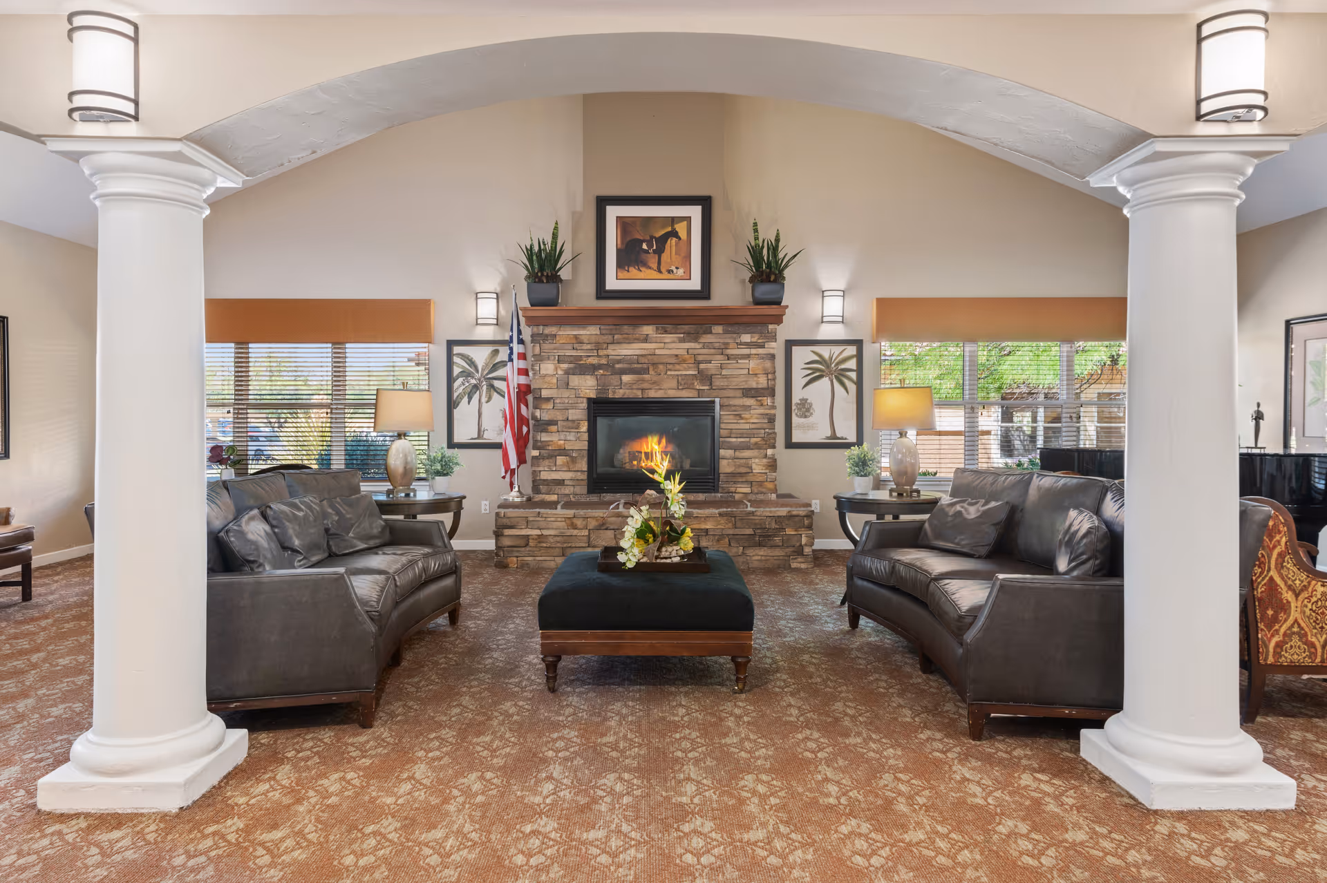 A cozy living room with two black leather sofas facing each other across a black ottoman with a floral arrangement. The room features a stone fireplace with a fire burning, flanked by two potted plants and framed artwork above. There are two large white columns framing the view, with windows on either side letting in natural light. The room has warm beige walls and carpeted flooring.
