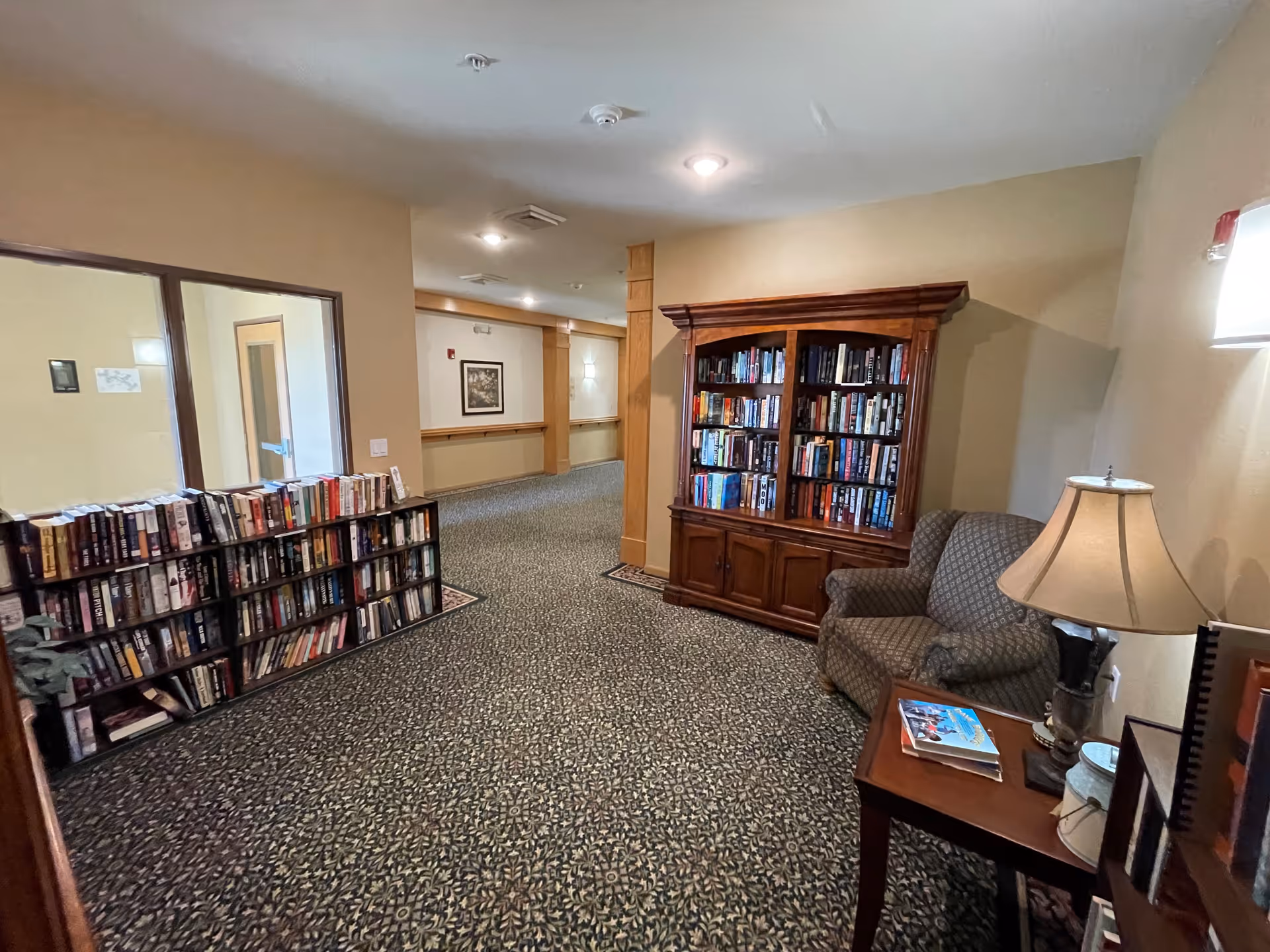 A cozy interior hallway area in a senior living facility featuring two wooden bookshelves filled with books, a patterned armchair, a small wooden side table with a lamp and magazines, and carpeted flooring with a decorative pattern.