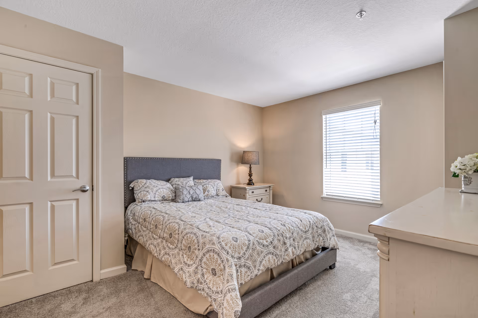 A bedroom with a neatly made bed featuring a patterned bedspread and multiple pillows. There is a nightstand with a lamp on the right side of the bed, a window with blinds on the far wall, a closed door on the left, and a dresser with a small flower arrangement on the right side of the image. The walls are painted beige and the floor is carpeted.
