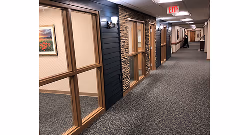 Carpeted interior hallway of a retirement facility with wood-and-stone accent walls, windows into rooms, wall sconces, and an EXIT sign.