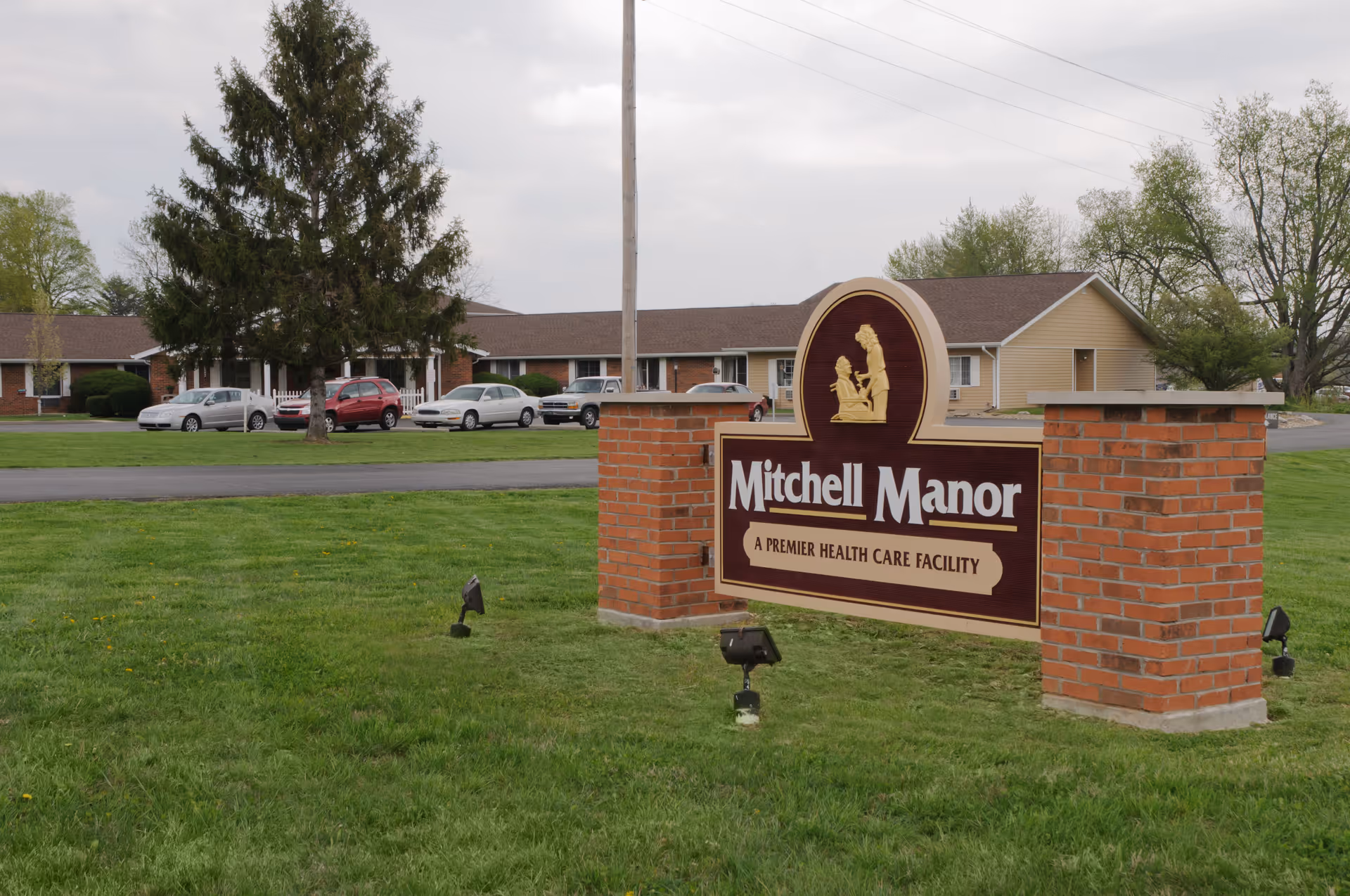 A brick sign reading "Mitchell Manor" on a grassy lawn in front of a single-story senior care building.