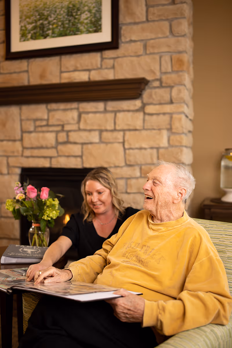 An elderly man and a caregiver sit in a cozy living room by a stone fireplace, smiling as they look through a photo album.
