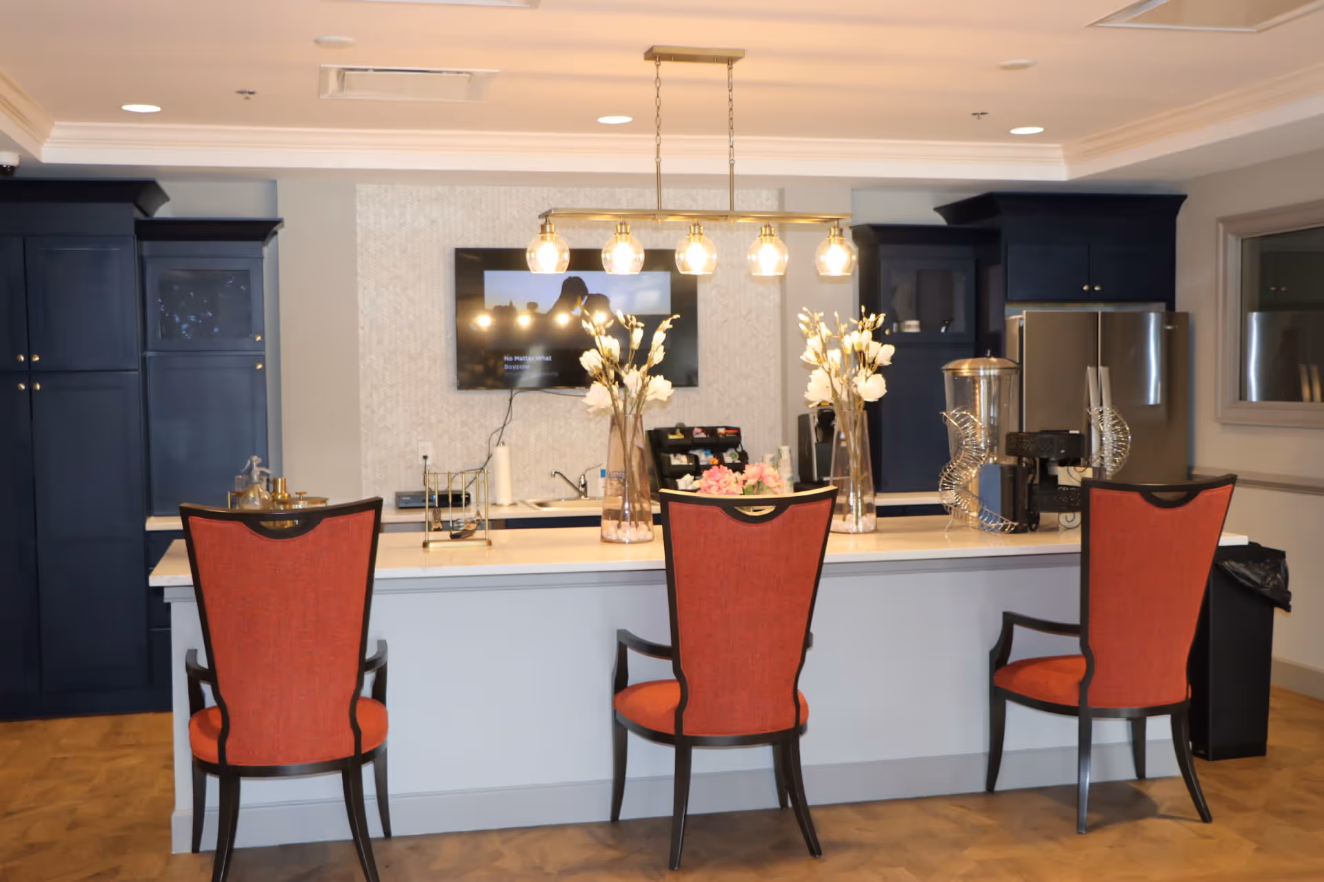 Interior view of a kitchen area with a white island countertop and three red cushioned chairs with dark wooden frames. The back wall features dark blue cabinets, a stainless steel refrigerator, a mounted flat-screen TV, and a sink. Two tall glass vases with white flowers are placed on the island. A modern light fixture with five bulbs hangs above the island.