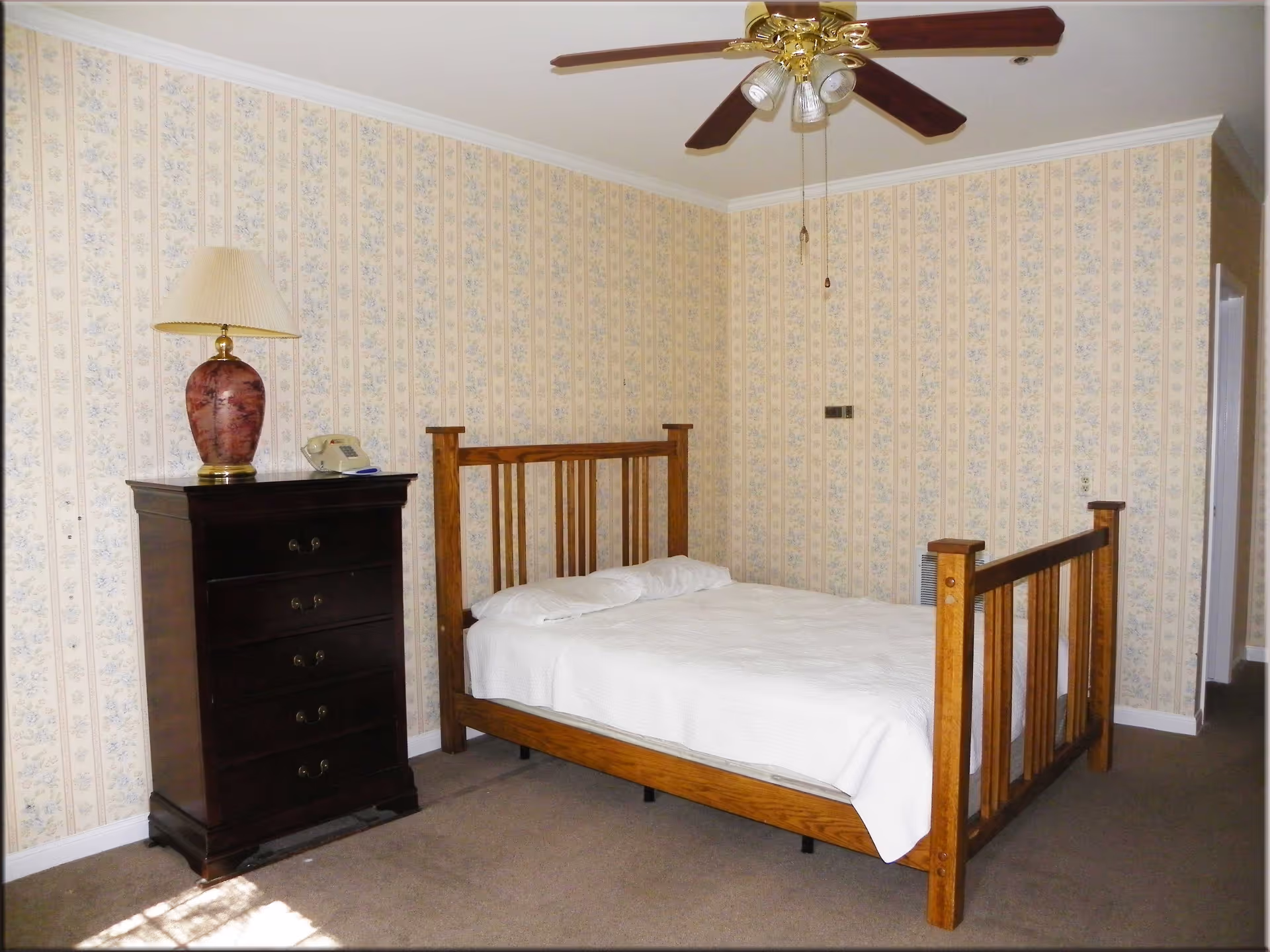 A bedroom with a wooden bed frame and white bedding, a dark wooden chest of drawers with a decorative lamp and a telephone on top, floral patterned wallpaper, a ceiling fan with lights, and a carpeted floor.