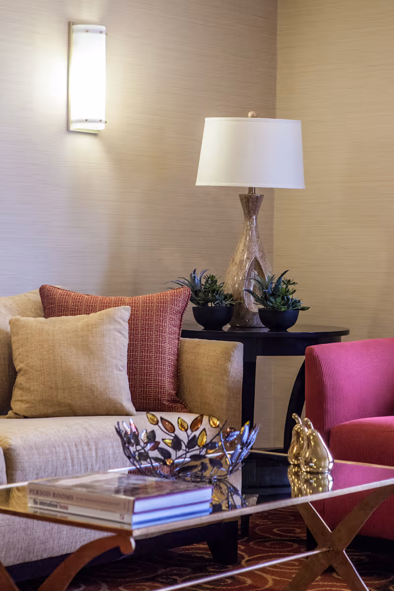 A cozy living room area with a beige sofa adorned with beige and red patterned pillows, a glass coffee table with decorative items and books, a side table with a stylish lamp and small potted plants, and a red armchair. The walls are light-colored with a wall-mounted light fixture.