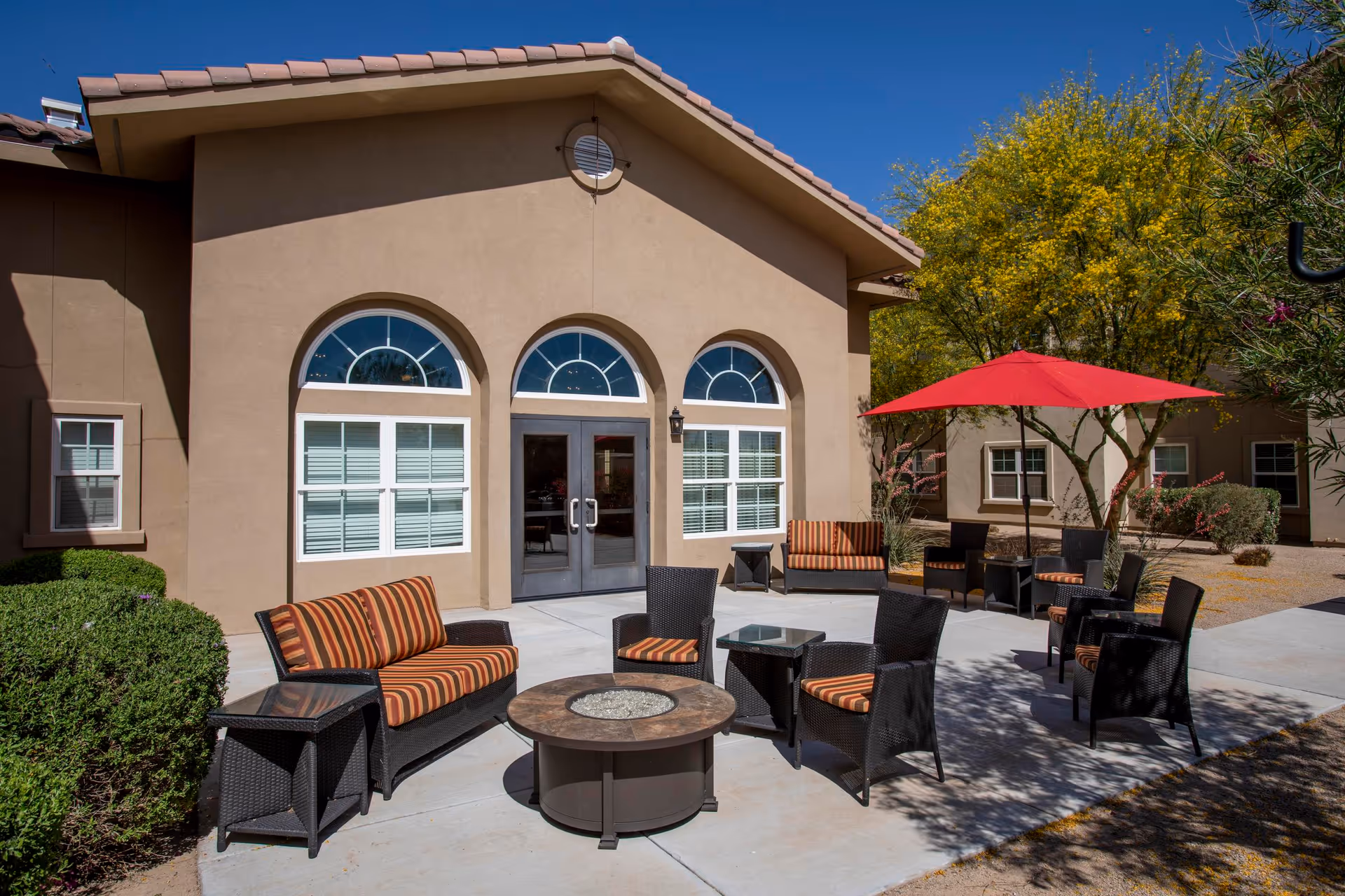 Outdoor patio area at Jaidon Manor assisted living with cushioned wicker chairs and sofas arranged around a circular fire pit table. There is a red umbrella providing shade over some seating. The building has beige stucco walls with three arched windows and double glass doors. Green bushes and trees with yellow leaves surround the patio under a clear blue sky.