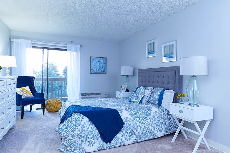 A bright and cozy bedroom featuring a large bed with blue and white patterned bedding and a navy blue throw blanket. The bed has a gray tufted headboard and is flanked by two white nightstands, each with a glass lamp and small decorative items. A navy blue armchair with a yellow pillow sits near a sliding glass door with sheer white curtains, leading to a balcony with a view of trees. The room has light gray walls adorned with framed artwork and a white dresser with a lamp on the left side.