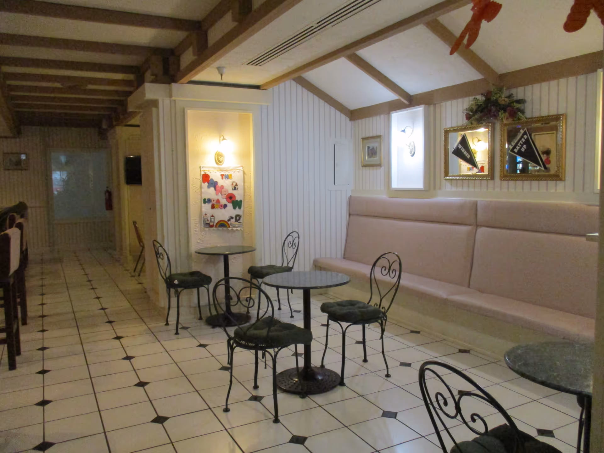 Small dining/lounge area with tiled floor, round bistro tables and wrought-iron chairs facing a long upholstered bench and framed mirrors.