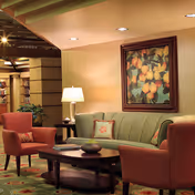 Comfortable lounge area with a green sofa, two coral armchairs, a coffee table, table lamp, and a framed fruit painting on the wall.