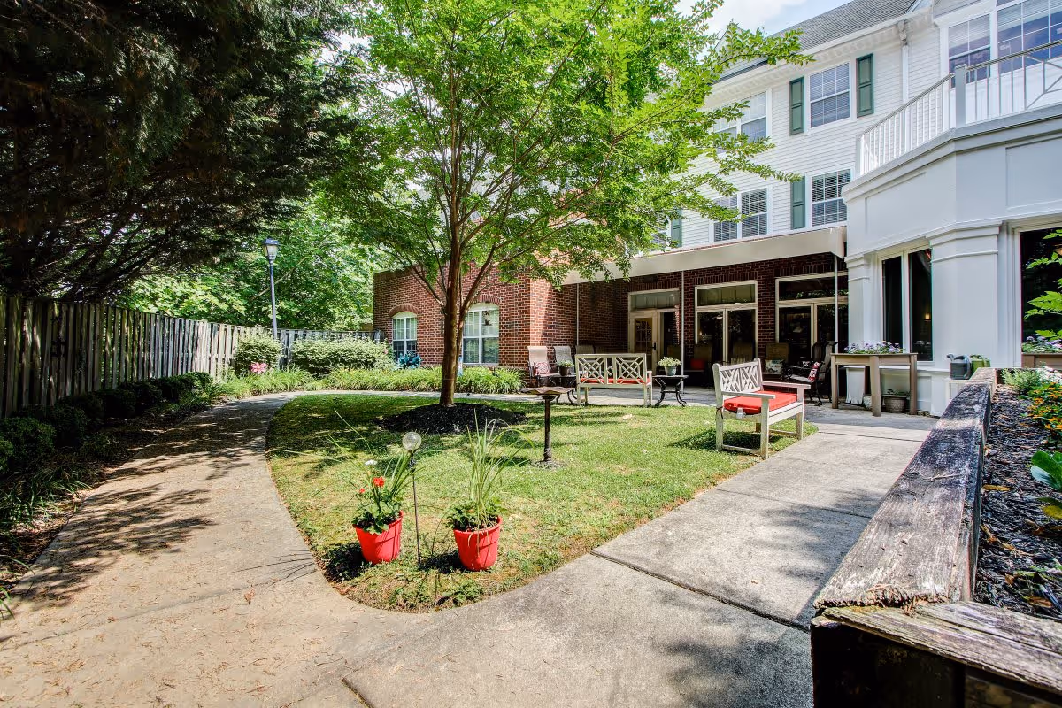 Outdoor courtyard area at Charter Senior Living of Woodholme Crossing featuring a paved walkway, green lawn with a tree, red potted plants, benches with red cushions, and a brick and white building in the background.