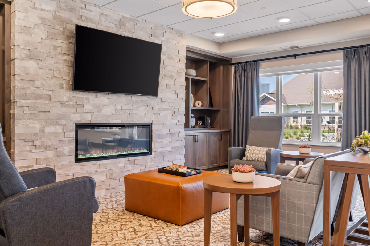 A cozy living room area with a stone accent wall featuring a mounted flat-screen TV above a modern electric fireplace. The room includes comfortable armchairs, a leather ottoman, wooden side tables with small potted plants, a built-in wooden cabinet with decorative items, and large windows with curtains allowing natural light to enter.