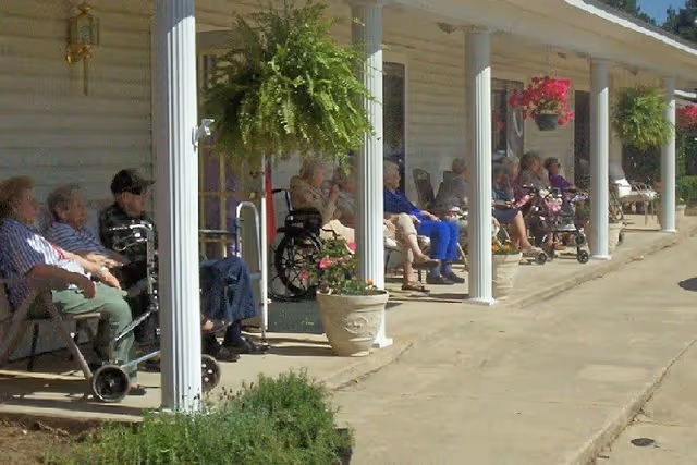 A group of elderly people sitting on chairs and wheelchairs along a covered porch outside a building, with hanging plants and potted flowers decorating the area.