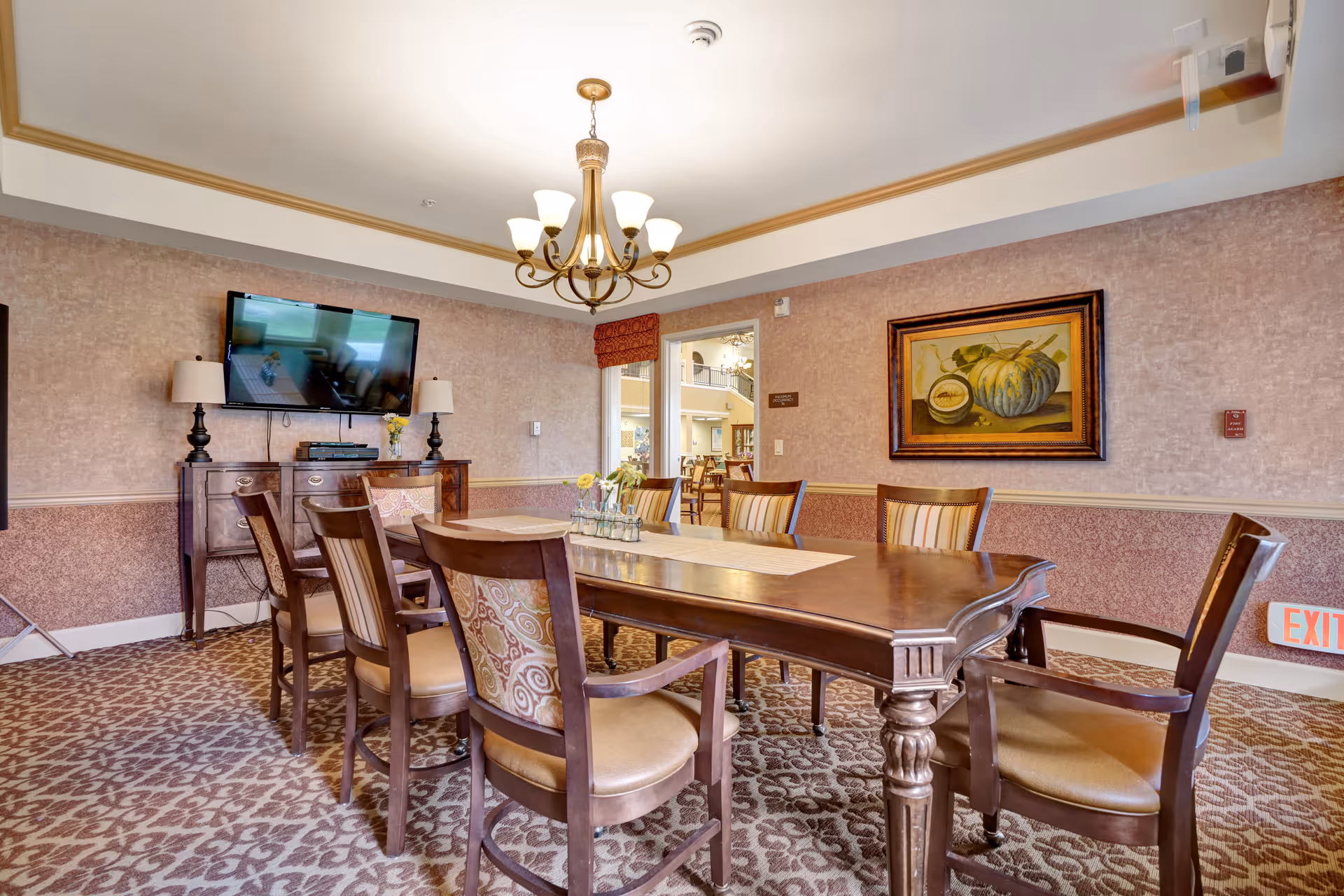 A dining room with a large wooden table surrounded by eight chairs with patterned upholstery. The room features a chandelier hanging from the ceiling, a flat-screen TV mounted on the wall above a wooden cabinet with two lamps, and a framed painting of pumpkins on the wall. The carpet has a floral pattern, and there is an exit sign near the floor on the right side.