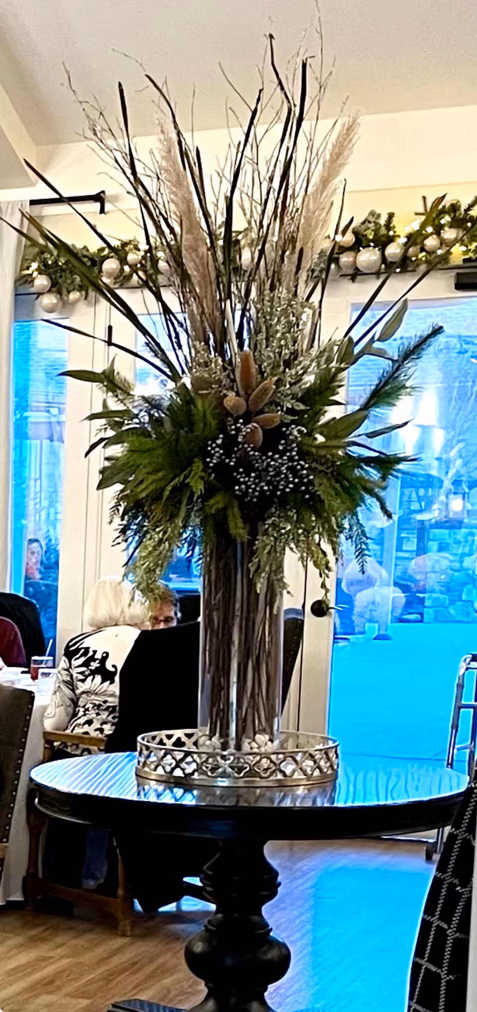 A tall floral arrangement with various green leaves, white flowers, and tall brown branches in a glass vase on a round wooden table inside a room. In the background, elderly people are seated at tables near large windows with a view of the outside.