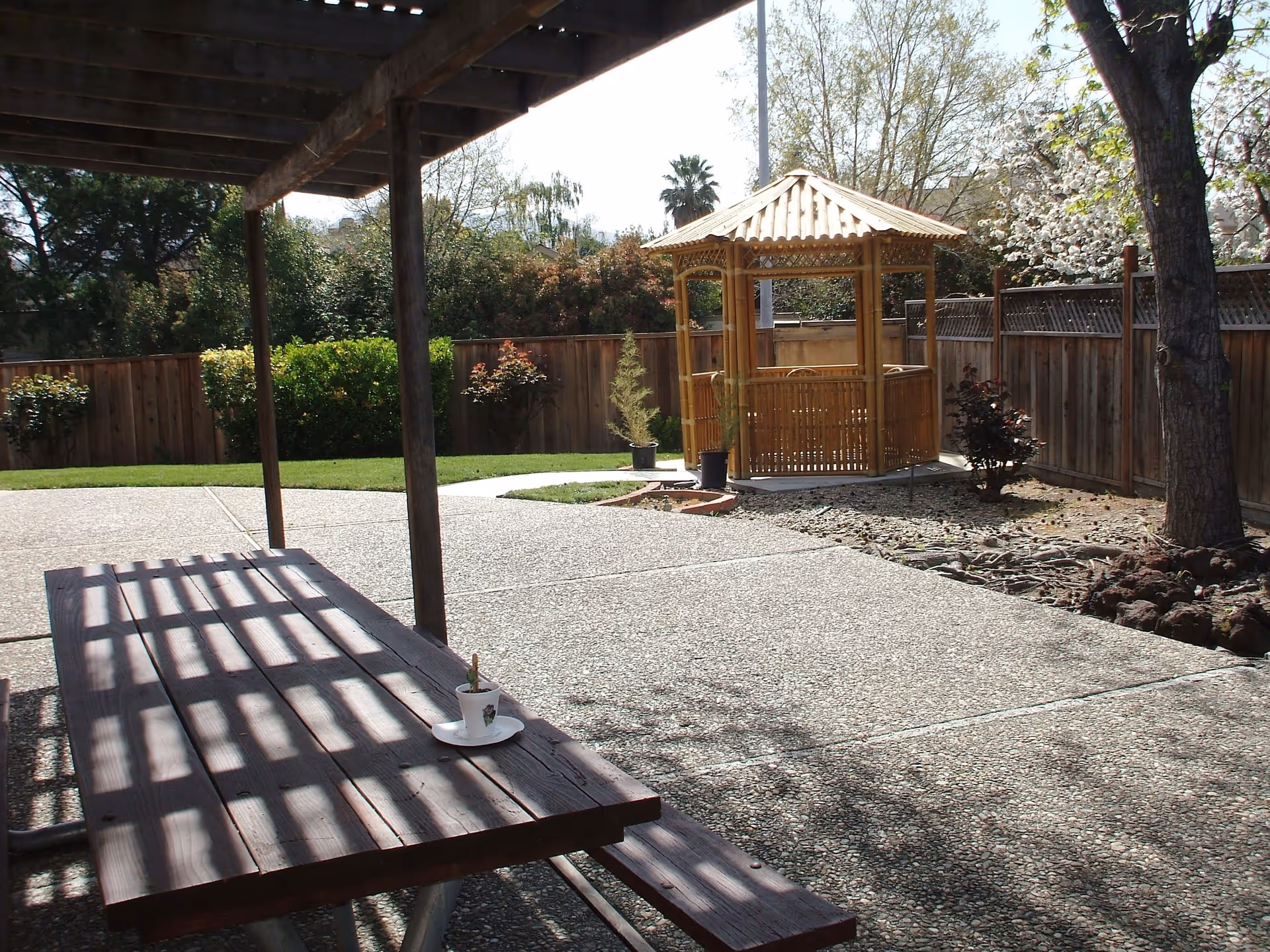 Outdoor patio area with a wooden picnic table under a pergola casting shadows, a concrete pathway, a small wooden gazebo, and surrounding greenery including trees and bushes enclosed by a wooden fence.